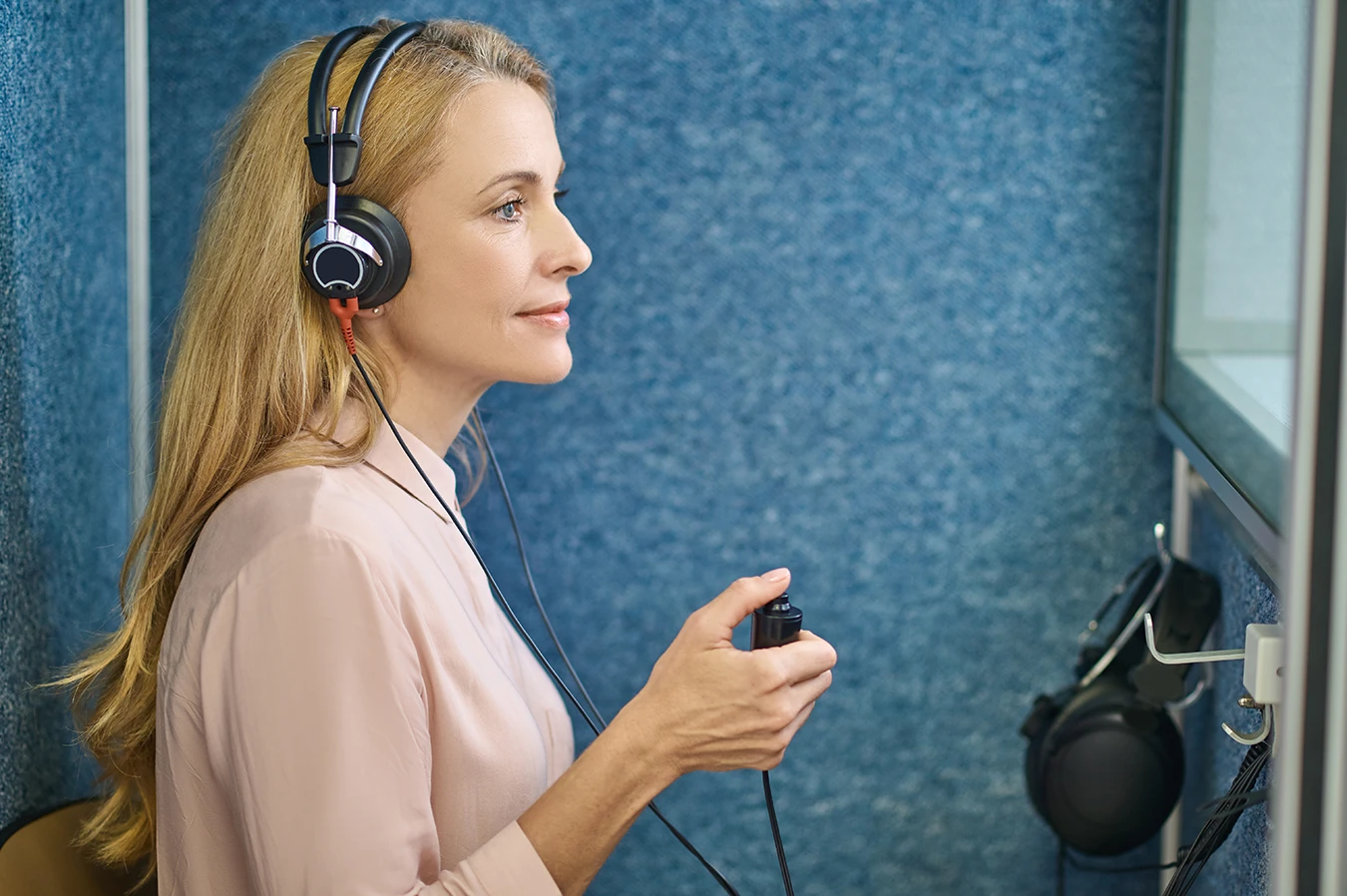 A middle aged woman with blonde hair sitting in a hearing test booth wearing over the ear headphones and holding an indicator button participating in a comprehensive hearing test at a hearing clinic